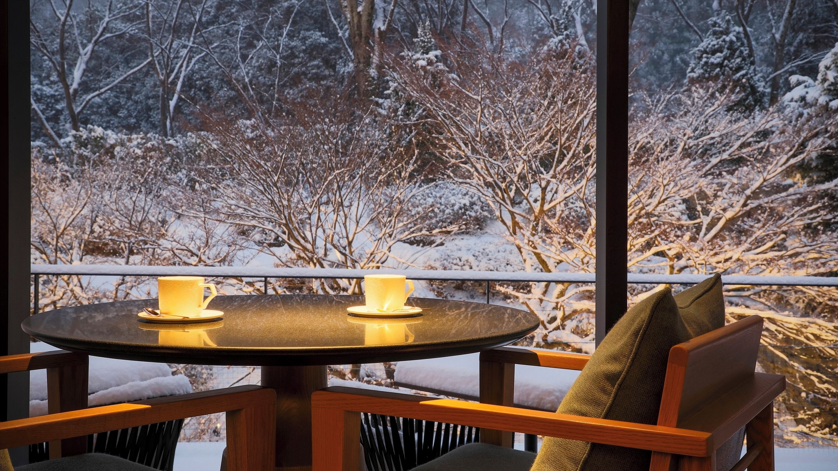 Une table ronde avec deux tasses et soucoupes jaunes, une chaise en bois avec un coussin, et une vue sur un paysage enneigé à travers de grandes fenêtres.