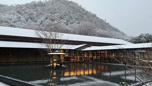 A traditional building with a reflective water feature in front, surrounded by snow-covered trees and a hill in the background.