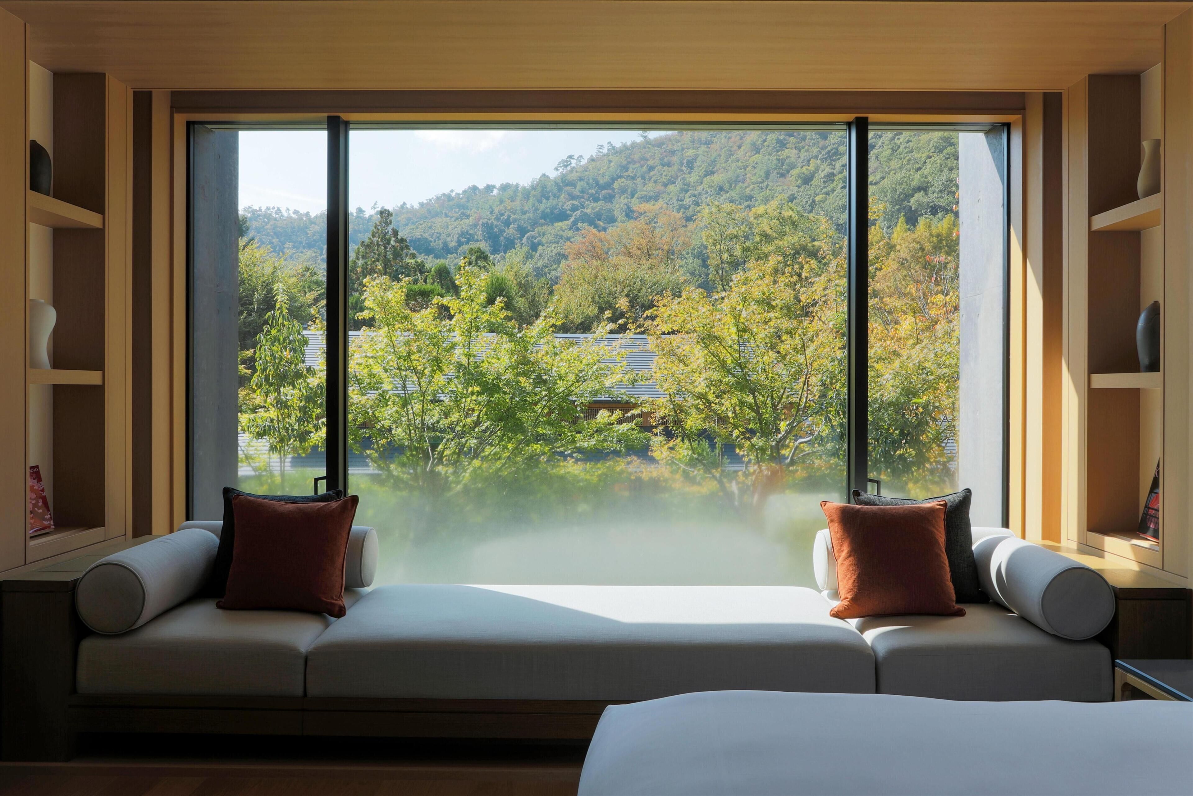 A modern room with a large window, a sofa with cushions, and a view of a forested area.