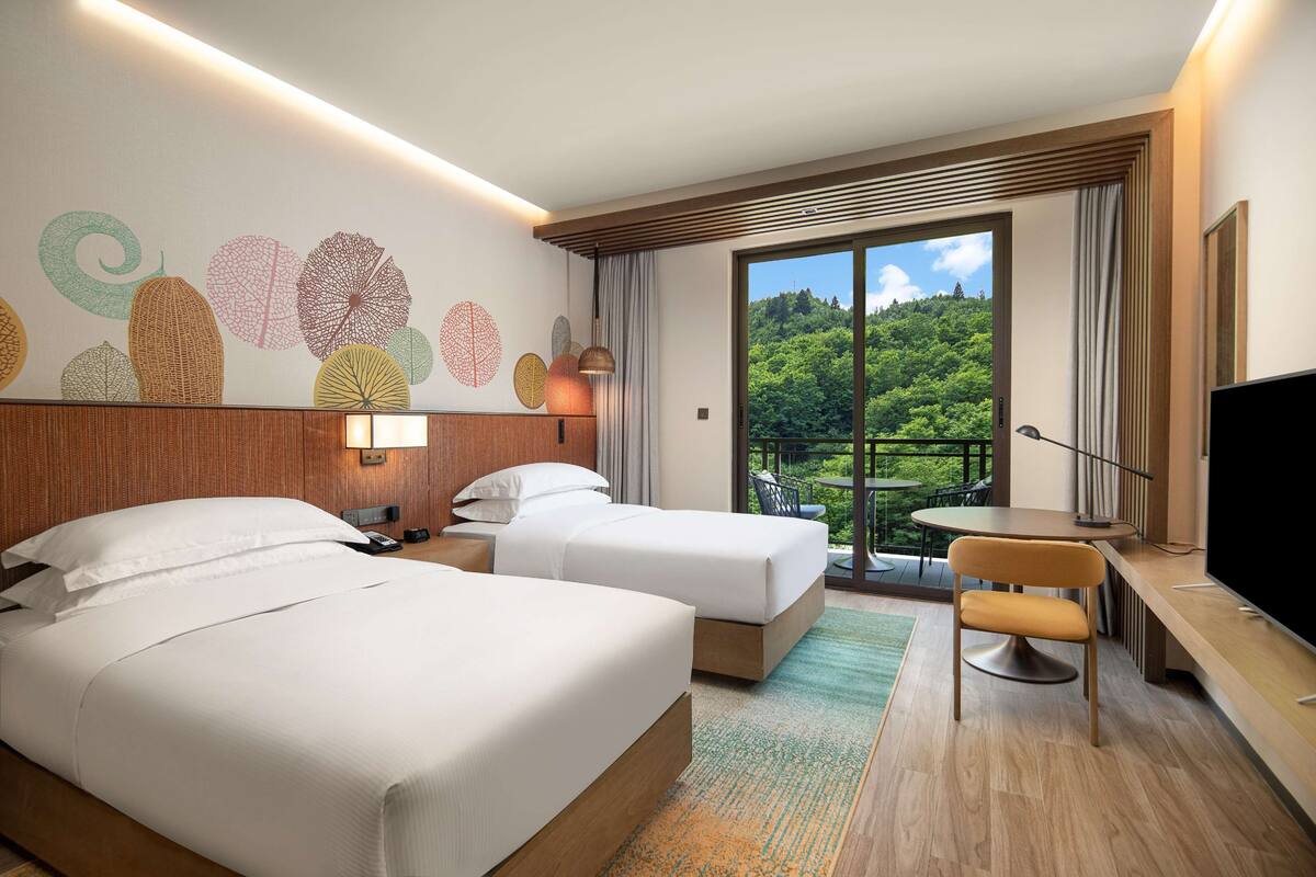 A hotel room with two beds, a desk, a chair, a TV, and a balcony with a view of greenery.