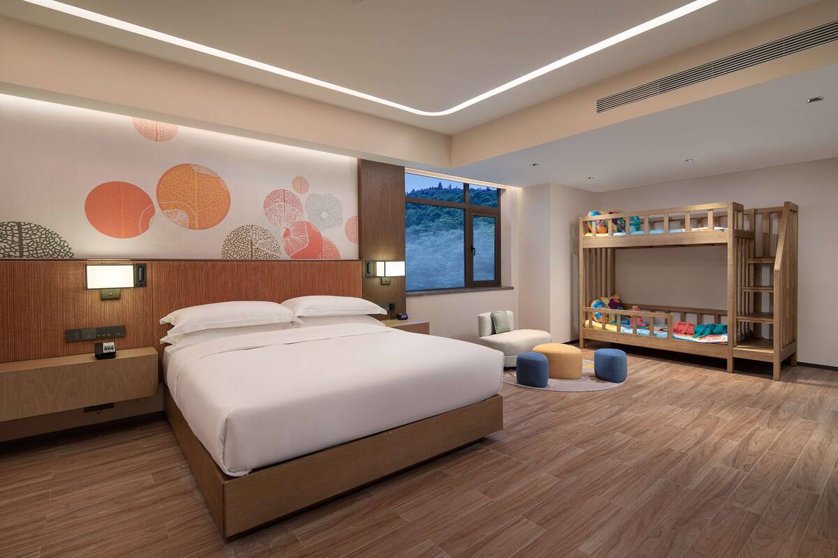 A modern hotel room with a large bed, a wooden bunk bed, a desk, and a wall with abstract art.