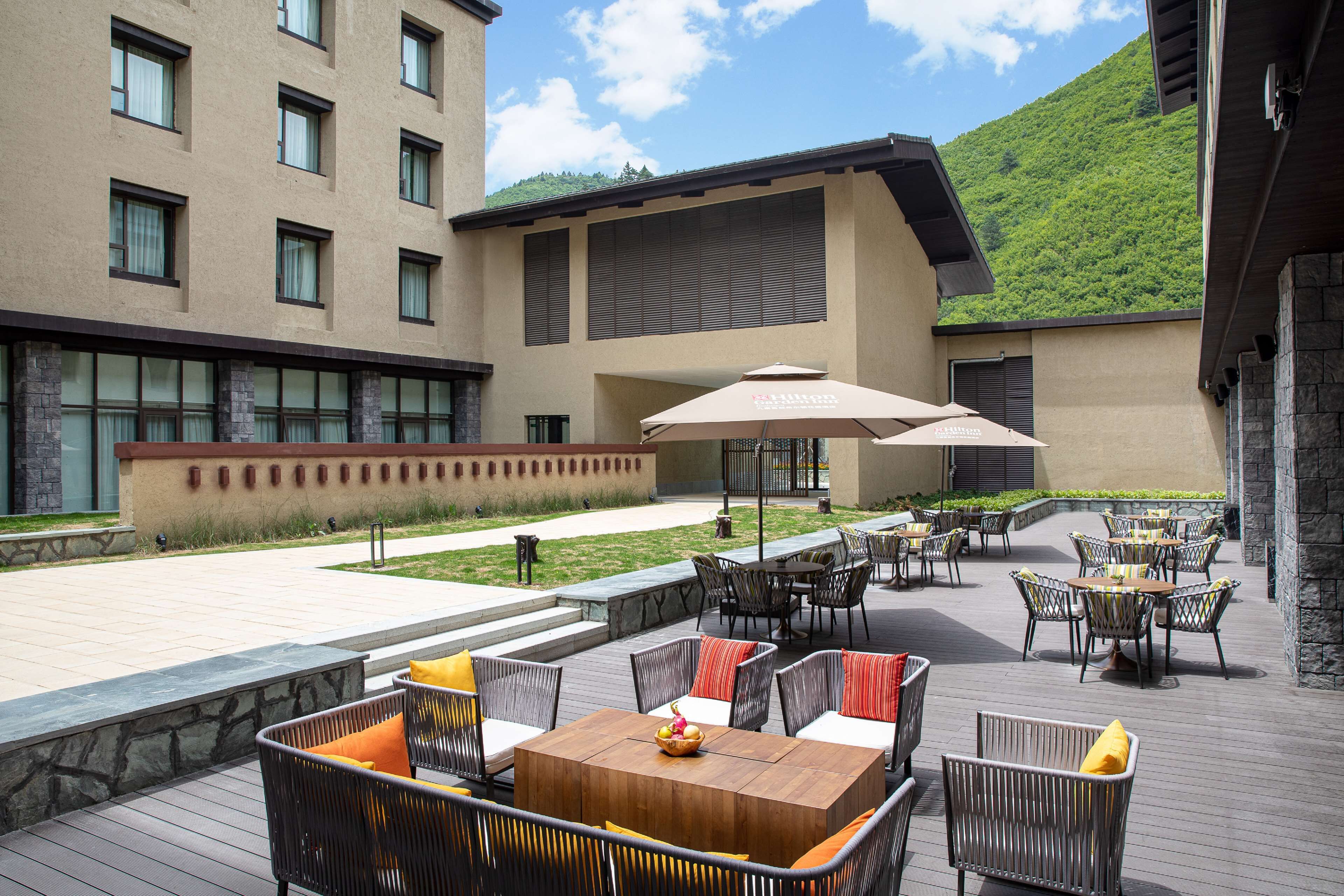 Un espace extérieur aménagé en terrasse, avec plusieurs tables et chaises, une plateforme en bois et un bâtiment doté de grandes fenêtres et d’une façade en pierre.
