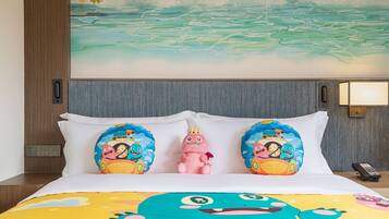 A hotel room with a bed featuring a colorful bedspread, two pillows with cartoon characters, a small pink toy, and a painting of a landscape on the wall.