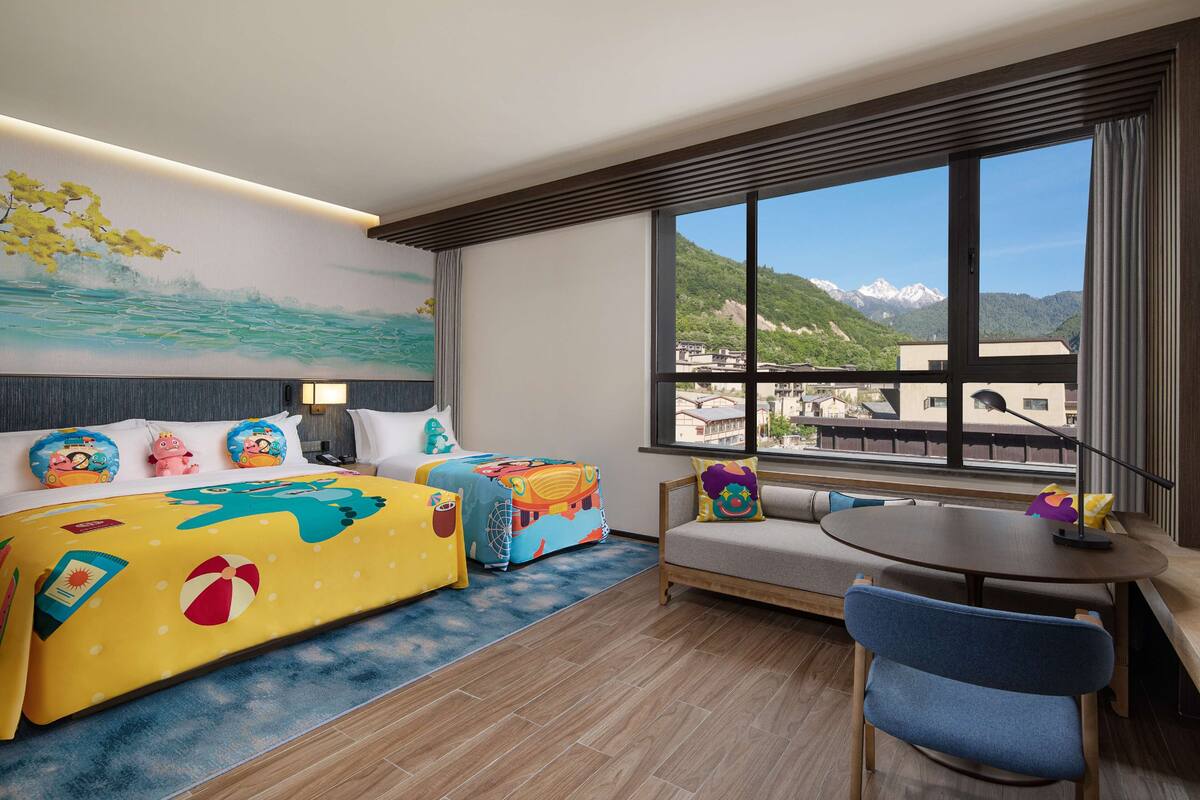 A hotel room with a bed, a sofa, a dining table, and a view of mountains.