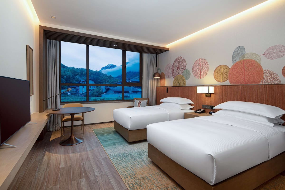 A hotel room with two beds, a TV, a desk, and a large window with a mountain view.