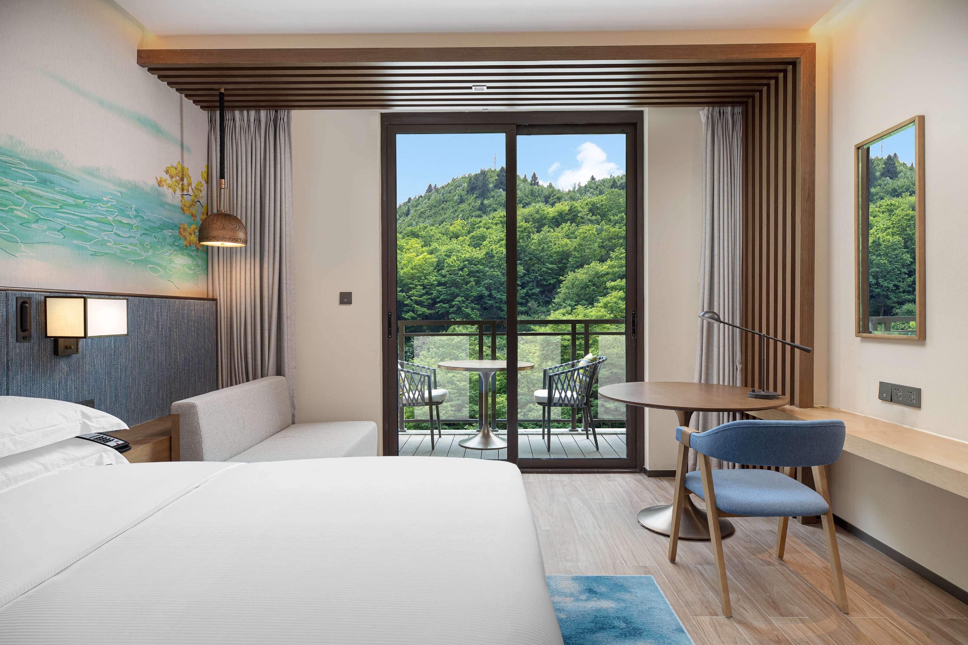A hotel room with a large bed, a desk, a chair, a balcony with a table and chairs, and a painting on the wall.
