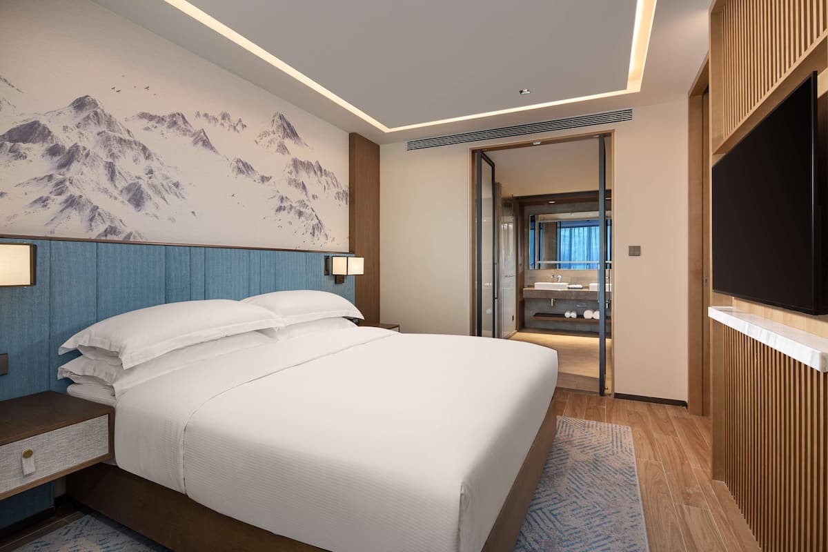 A modern hotel room with a large bed, a mounted TV, and a bathroom visible through an open door.