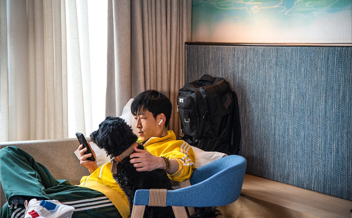 A person sitting on a blue chair in a room with a large window, a wall with a textured pattern, and a black backpack on the floor.