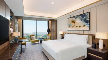 A hotel room with a large bed, a desk, a chair, and a TV. There is a large window with a view of the city.