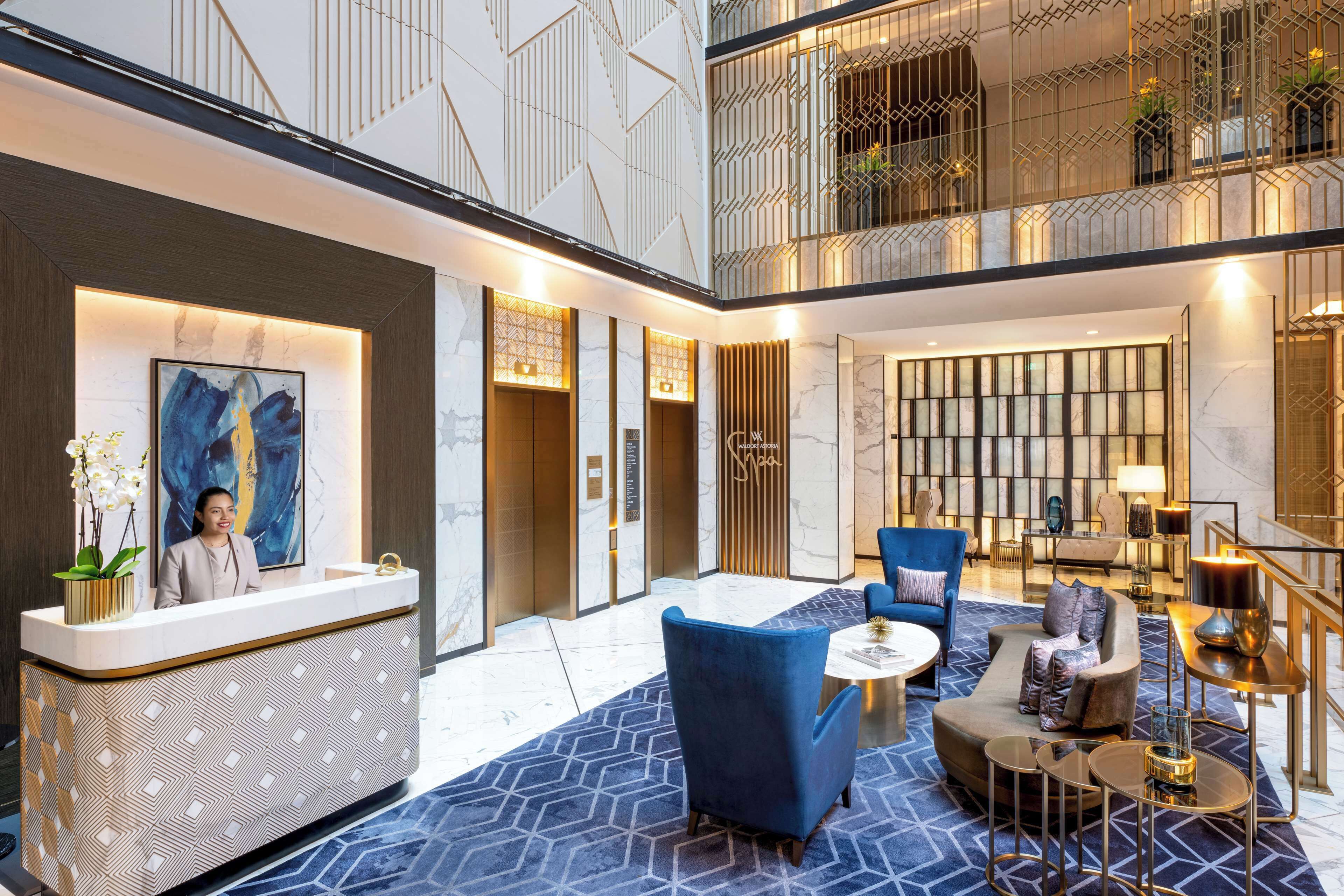 A modern hotel lobby with a reception desk, seating area, and a staircase leading to an upper level.