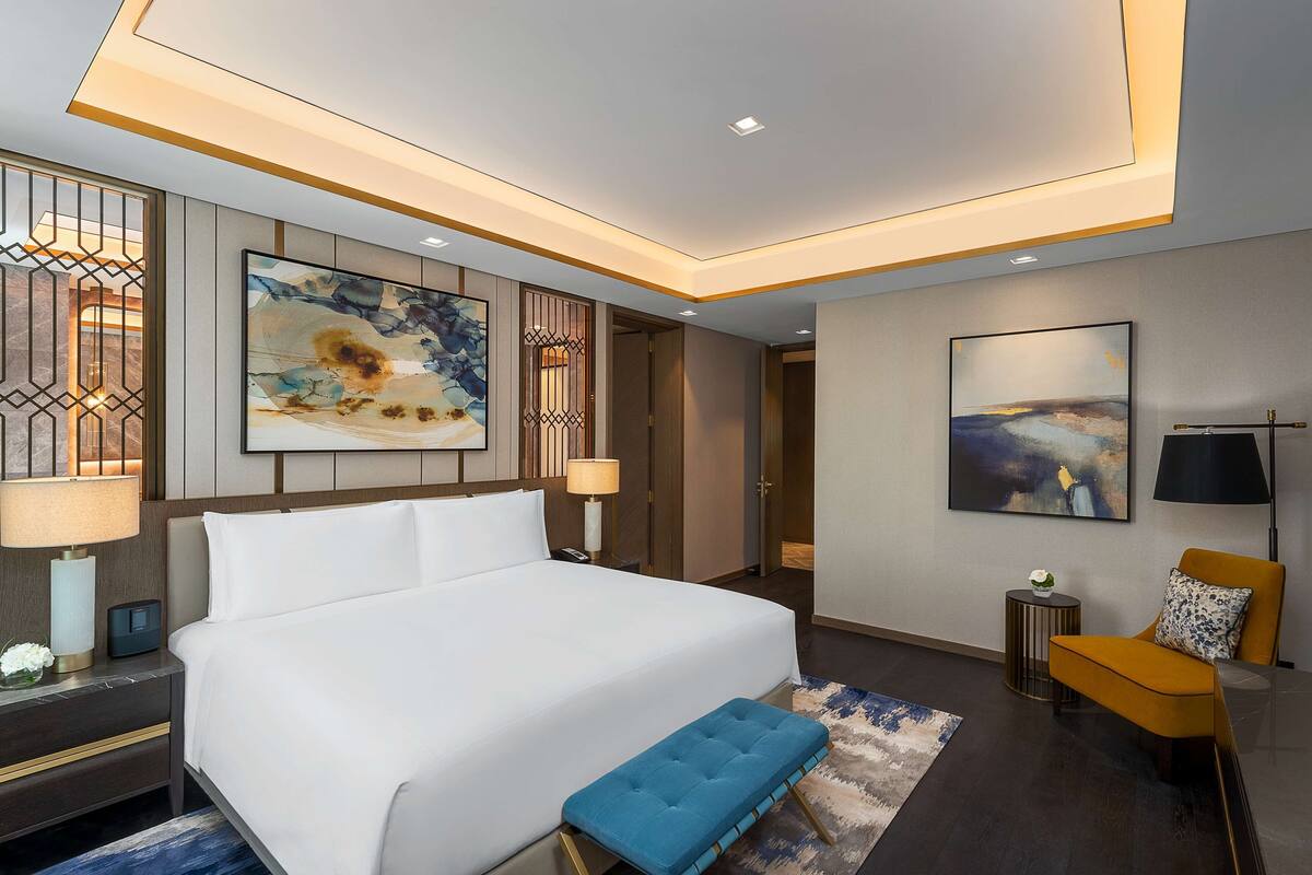 Una habitación de hotel moderna con una cama grande, un banco azul, una mesita auxiliar pequeña y una silla.