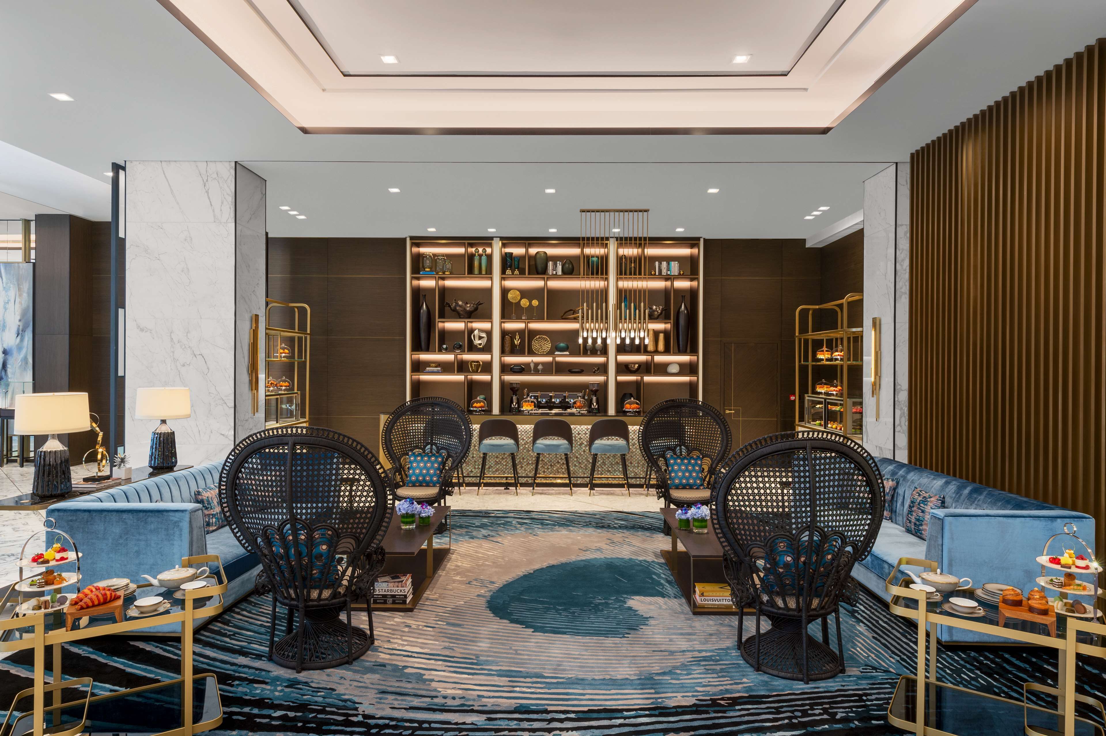 Un moderno vestíbulo de hotel con un sofá azul, sillas de mimbre, un mostrador de recepción de mármol y una estantería llena de objetos decorativos.