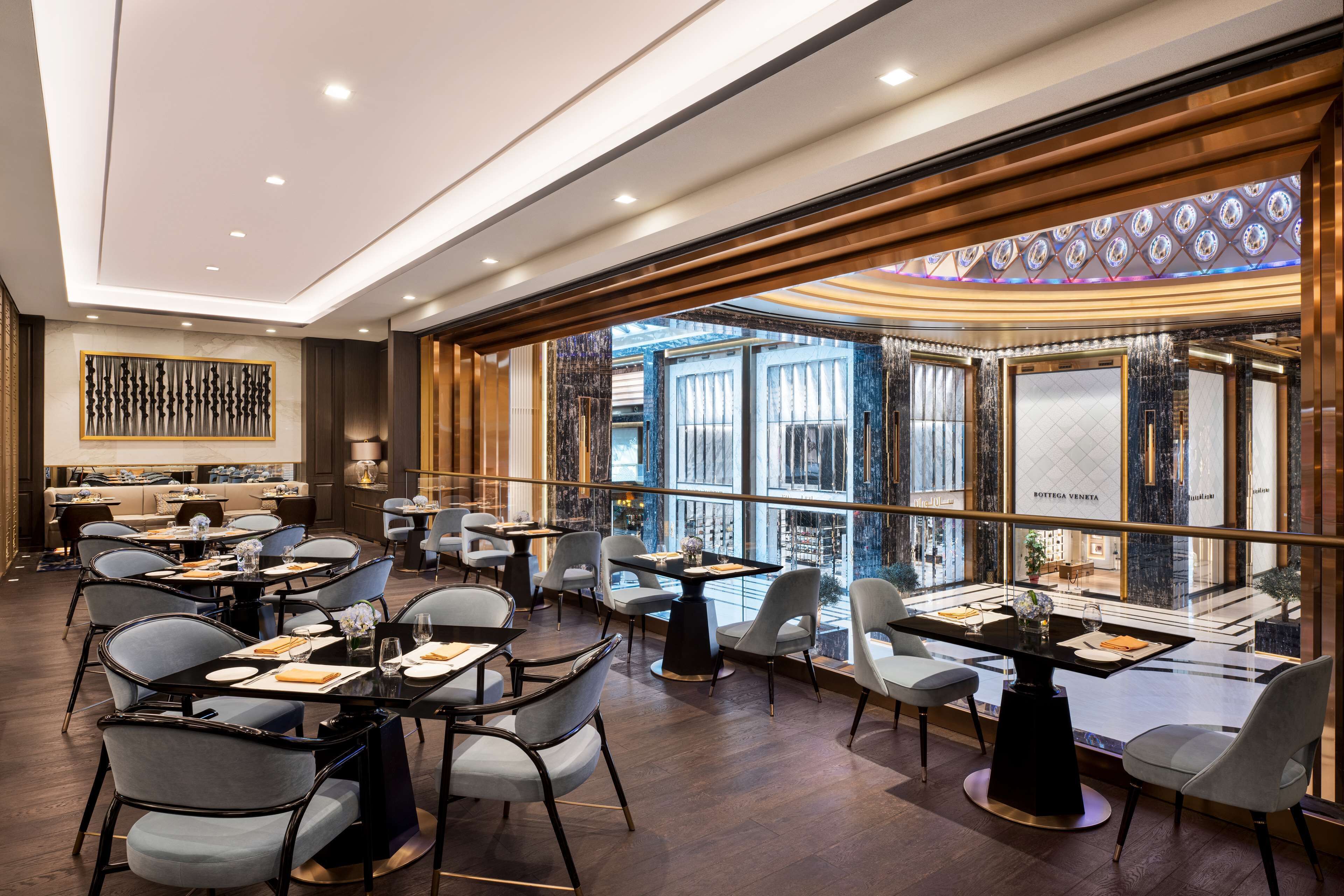 A modern restaurant with multiple dining tables and chairs, a decorative wall, and large windows offering a city view.