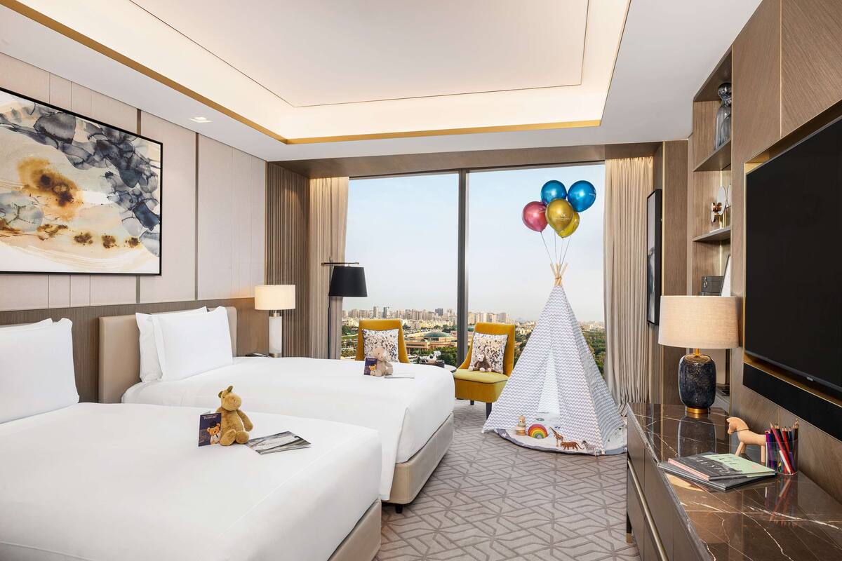 Habitación de hotel con dos camas, un ventanal grande con vistas a la ciudad, televisión y una pequeña tienda de campaña con globos.