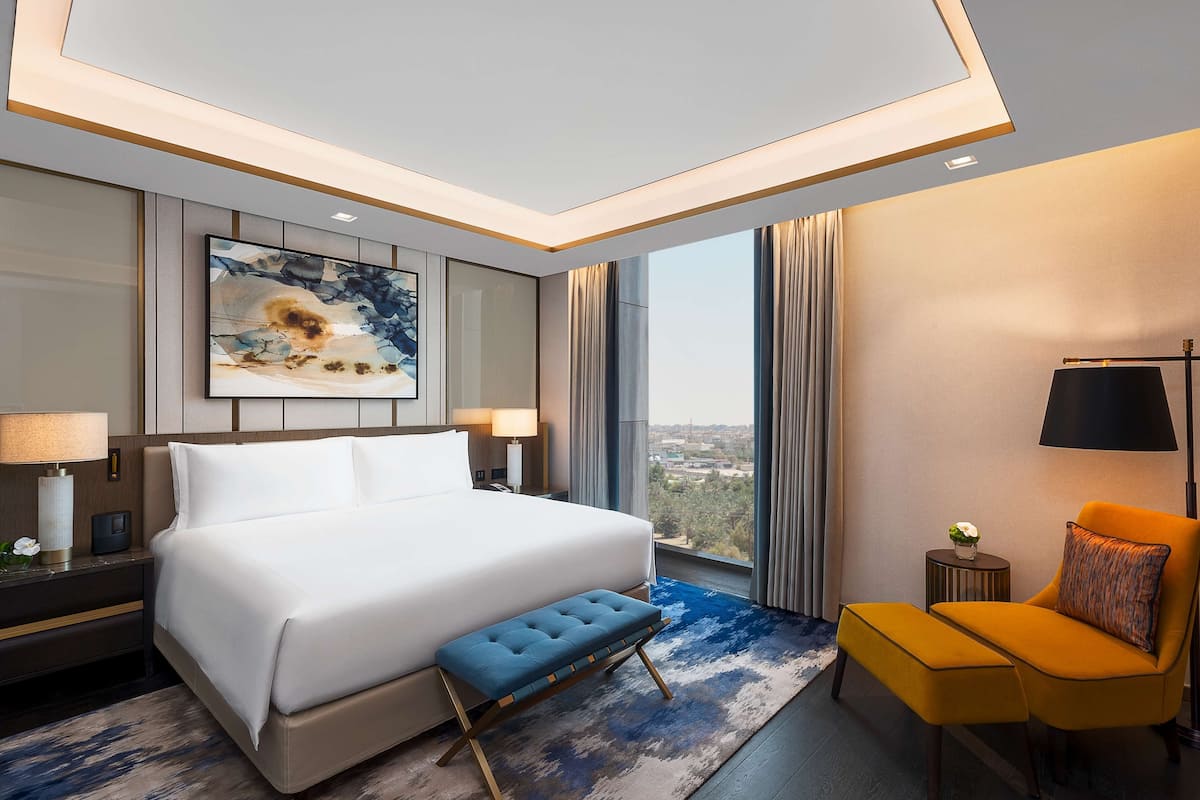 Una habitación de hotel moderna con una cama grande, un banco, una silla, una mesita de noche, una lámpara y un cuadro en la pared.