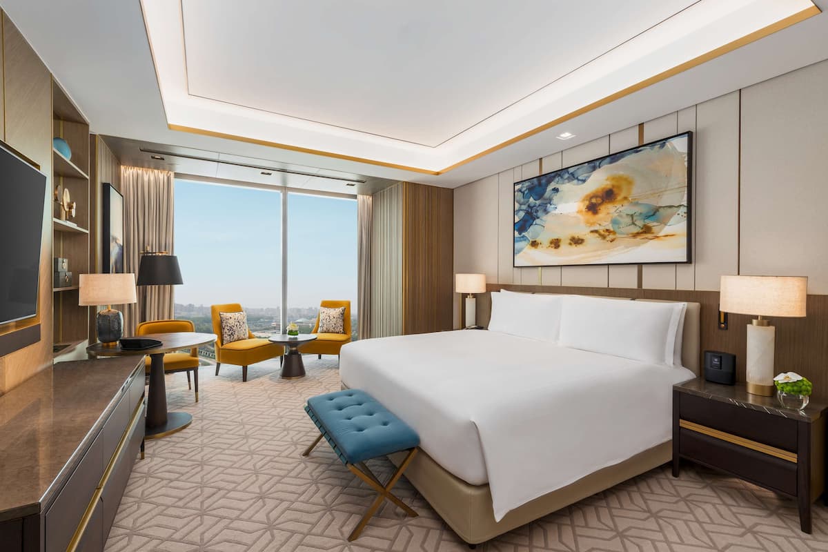 Habitación de hotel con una cama grande, un escritorio con televisor de pantalla plana, una zona de comedor con sillas amarillas y un ventanal con vistas a la ciudad.