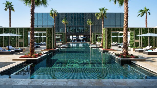 Un área de piscina moderna con sillas de descanso, palmeras y un edificio de cristal al fondo.