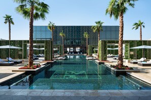 Un área de piscina moderna con sillas de descanso, palmeras y un edificio de cristal al fondo.