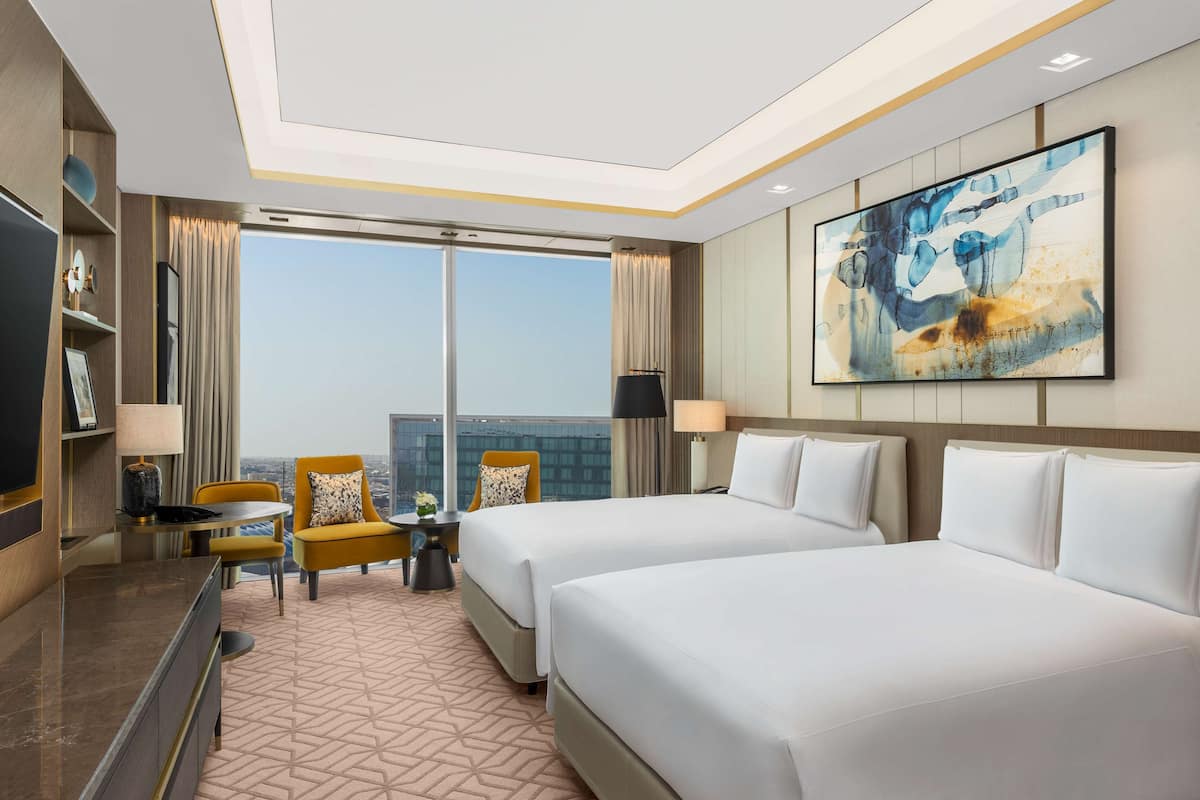 Habitación de hotel con dos camas, un escritorio, una silla, un televisor y un ventanal con vistas a la ciudad.