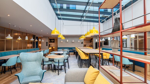 A modern dining area with high ceilings, yellow pendant lights, and a mix of seating options including tables and booths. - Hampton by Hilton Canterbury (Canterbury)