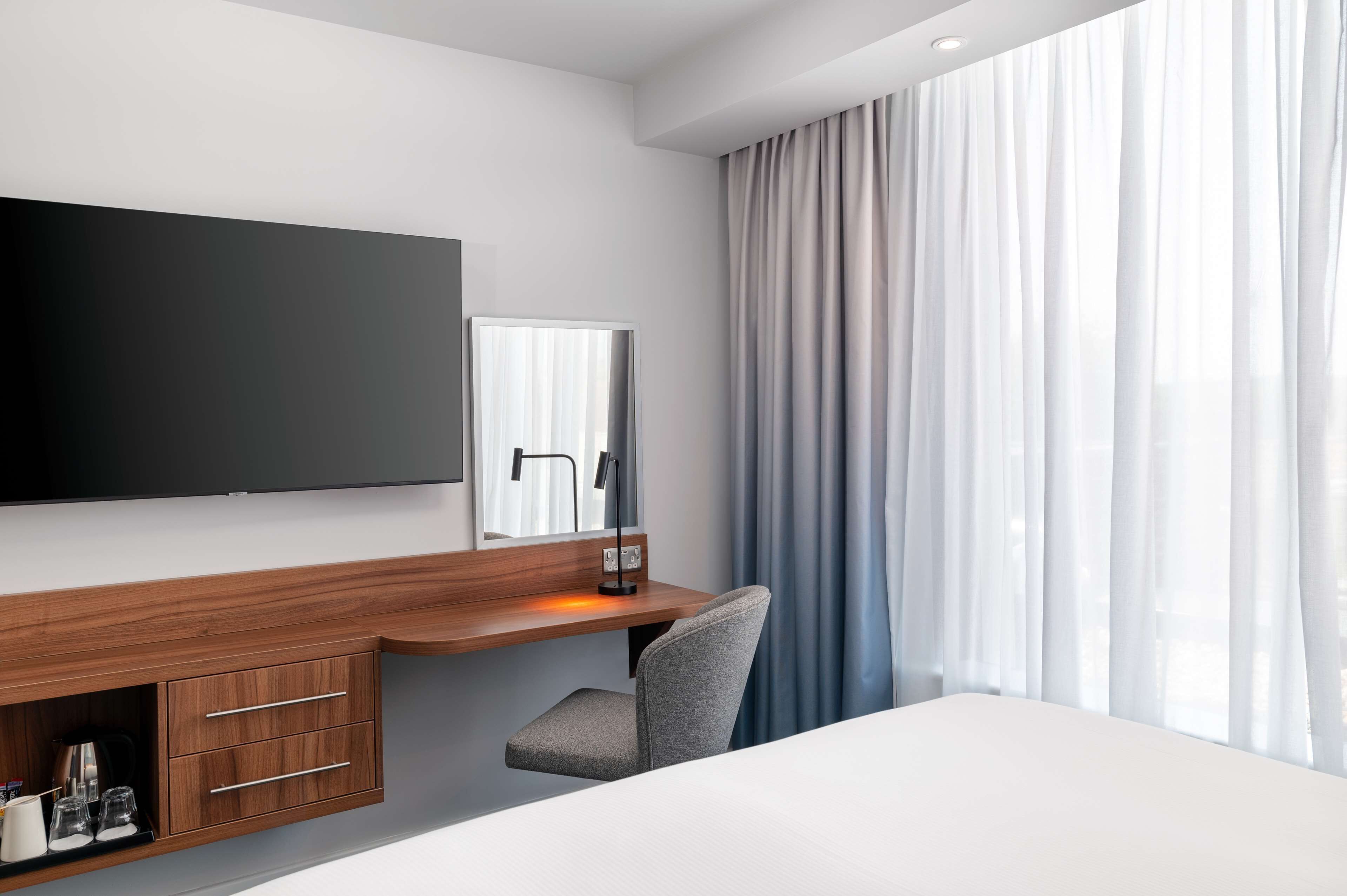 A modern hotel room with a flat-screen TV, a desk with a chair, a large window with curtains, and a bed.