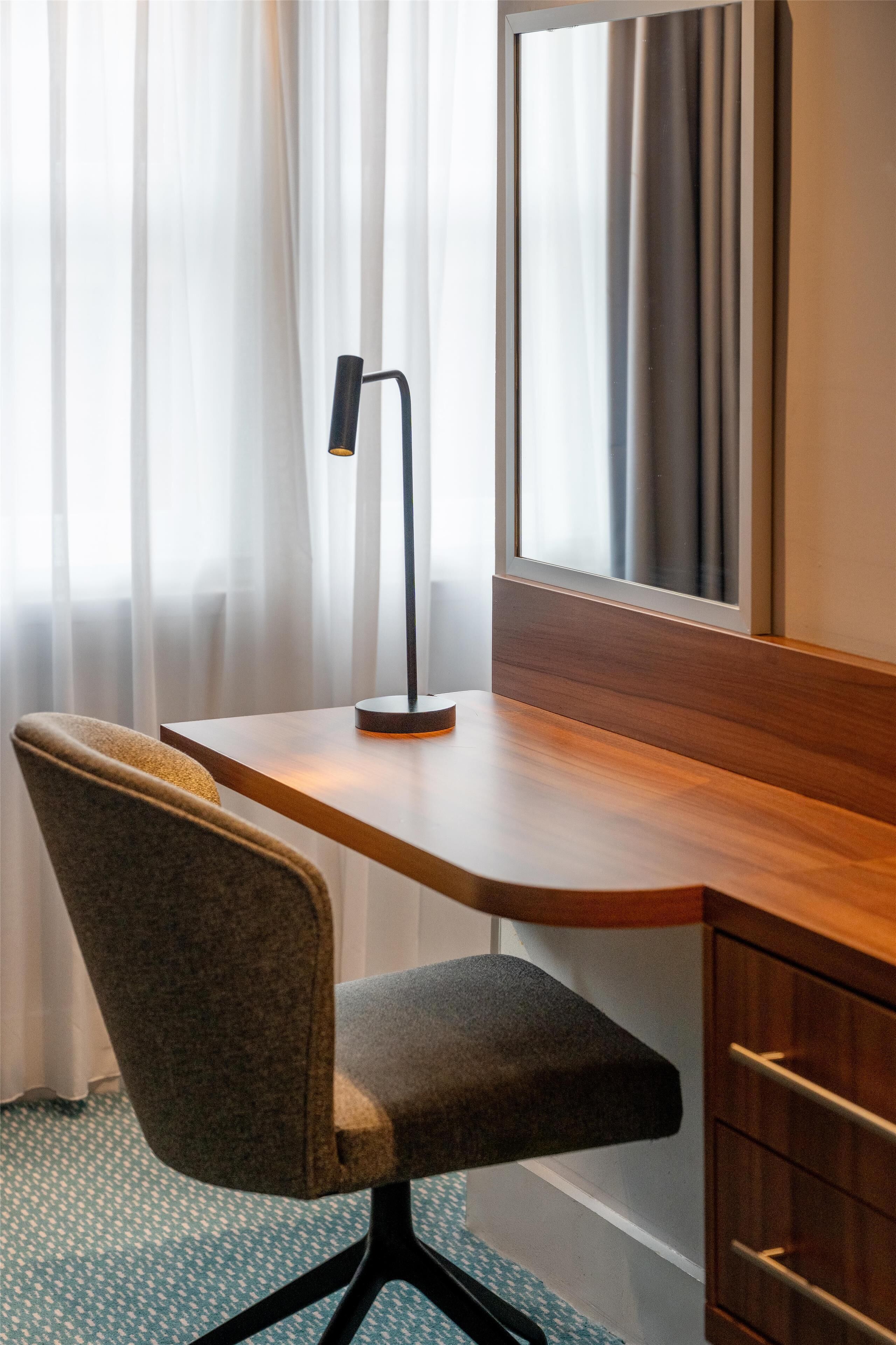 Une chambre d’hôtel avec un bureau en bois, une lampe de bureau moderne, une chaise, un grand miroir et une fenêtre avec des rideaux légers.