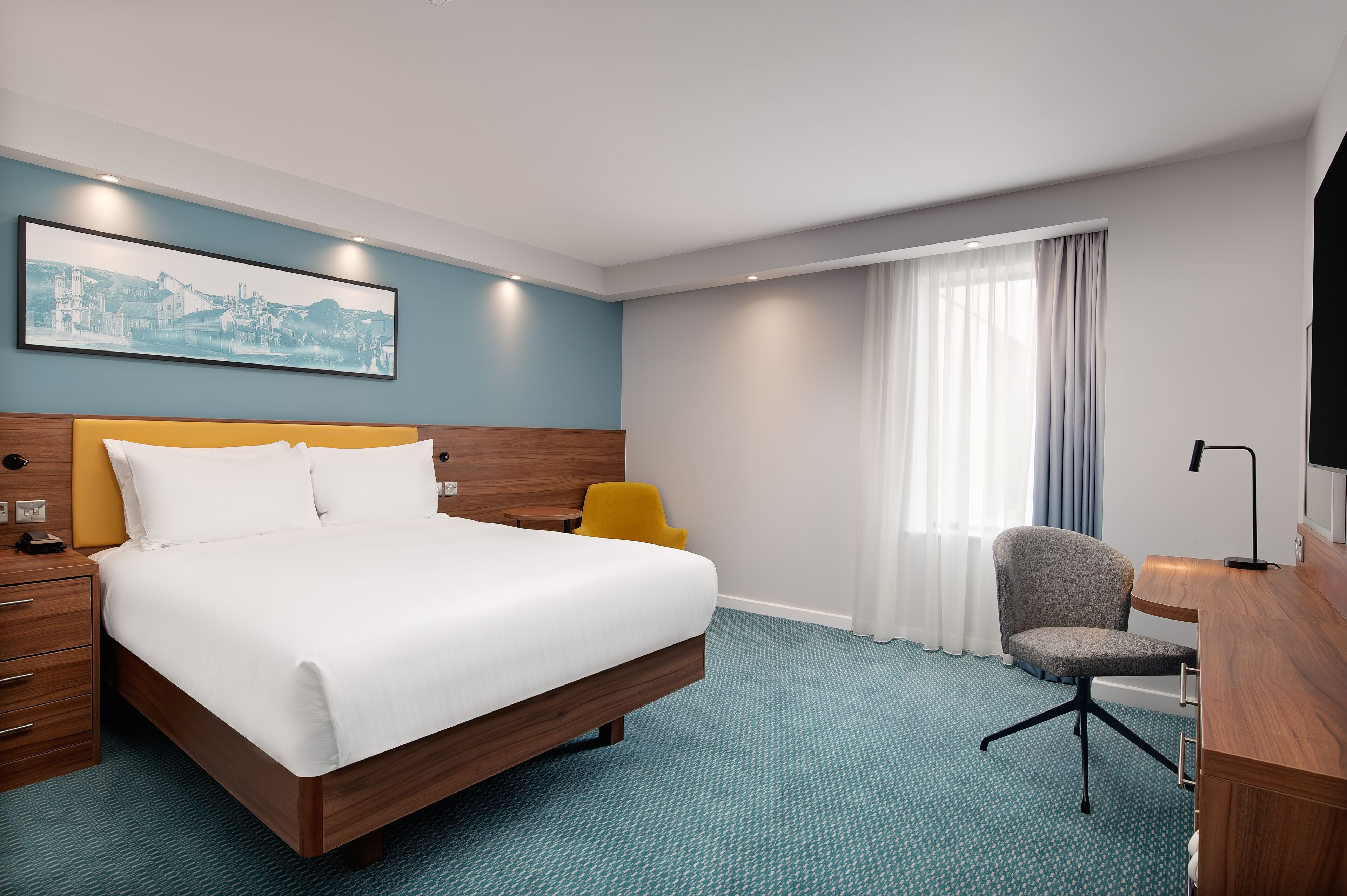 A hotel room with a bed, a desk, a chair, a nightstand, a lamp, and a window with curtains.