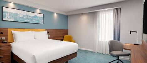 A hotel room with a bed, a desk, a chair, a nightstand, a lamp, and a window with curtains.