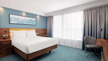 A hotel room with a large bed, a desk with a chair, a nightstand, a framed picture on the wall, and a window with curtains.
