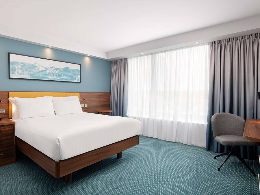 A hotel room with a large bed, a desk with a chair, a nightstand, a framed picture on the wall, and a window with curtains.