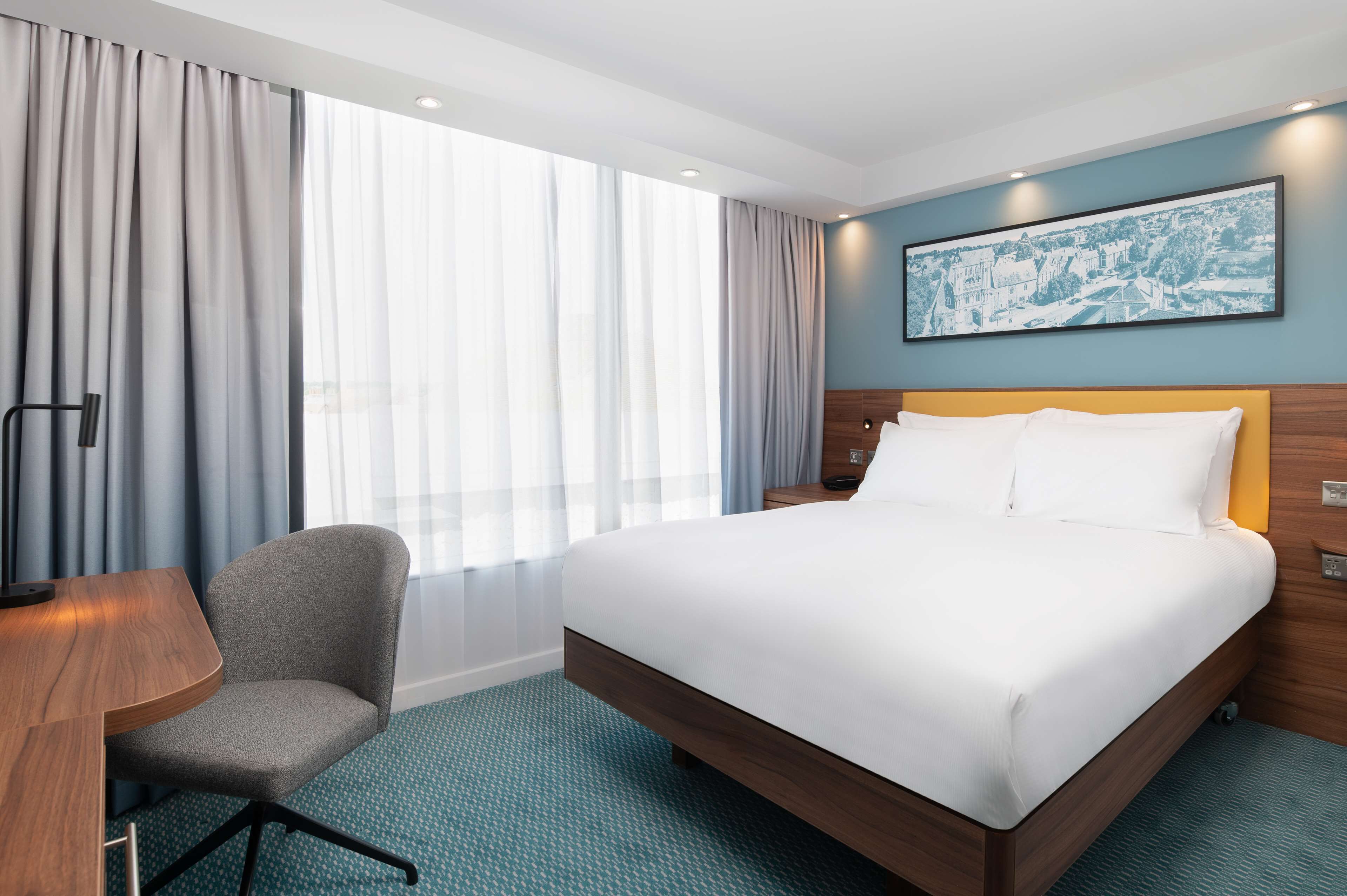 A hotel room with a large bed, a desk, and a chair. There is a window with curtains, a wall-mounted painting, and a carpeted floor.