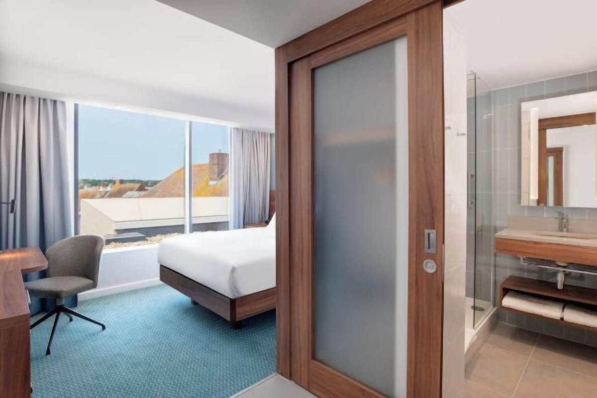 A hotel room with a bed, a desk, a chair, a bathroom with a mirror, and a view of the outside.