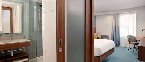 Quarto de hotel com chuveiro em cabine de vidro, toucador de madeira, cama, escrivaninha e cadeira.