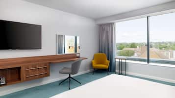 A hotel room with a large bed, a desk with a flat-screen TV, a chair, and a large window with a city view.