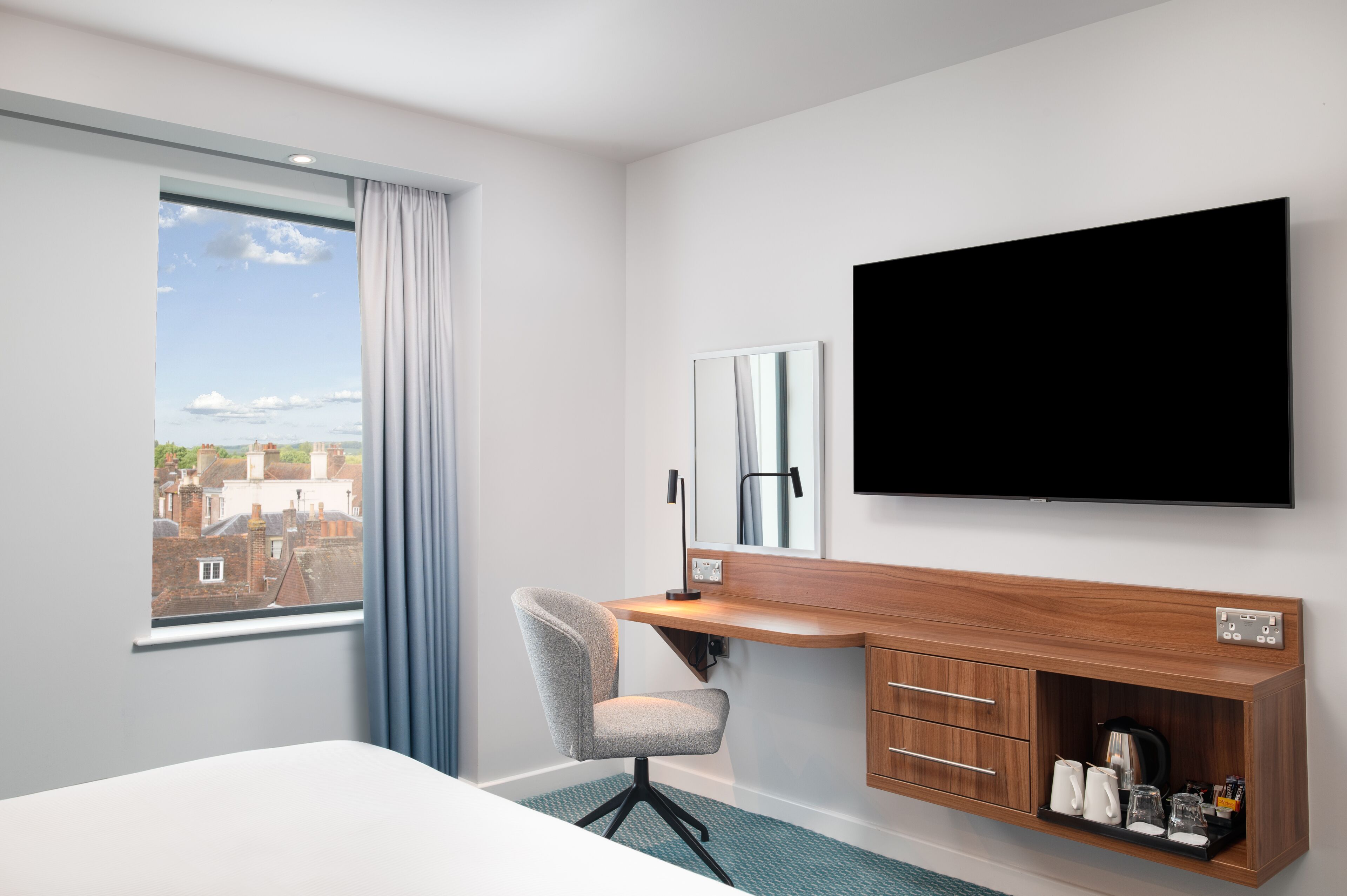 Habitación de hotel con cama, escritorio y silla, televisor de pantalla plana, espejo y vista a la ciudad a través de una ventana.
