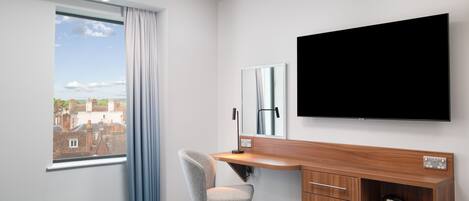 Habitación de hotel con cama, escritorio y silla, televisor de pantalla plana, espejo y vista a la ciudad a través de una ventana.