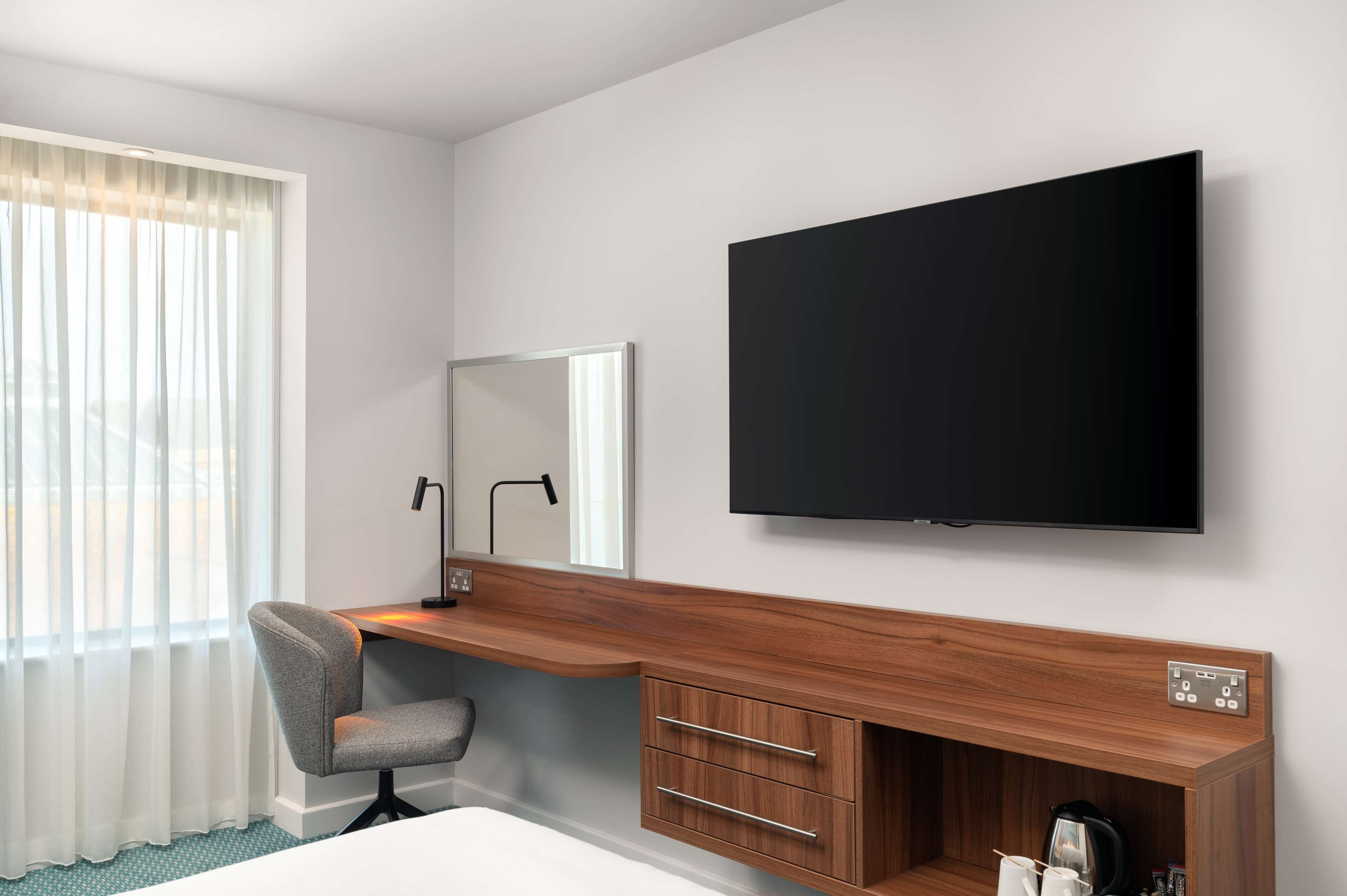 Habitación de hotel con escritorio de madera, televisor de pantalla plana, una silla, cama y ventana con cortinas.