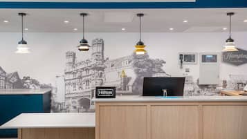 A hotel reception desk with a "Hilton Honors" sign, a wall-mounted phone, a digital display, and a "Hampton by Hilton" sign.