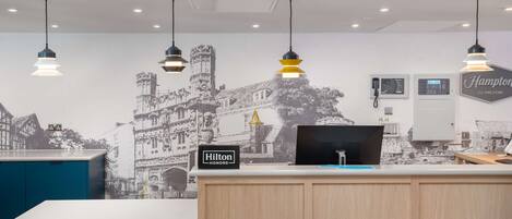 Un mostrador de recepción de hotel con un cartel de "Hilton Honors", un teléfono de pared, una pantalla digital y un cartel de "Hampton by Hilton".
