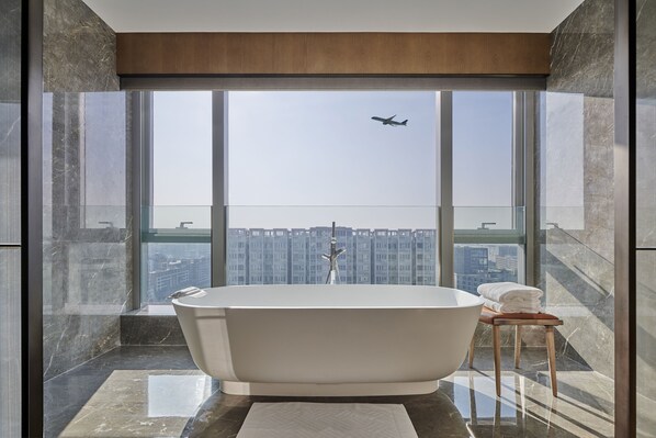Un baño moderno con una bañera independiente, un taburete de madera y un amplio ventanal con vistas a la ciudad.