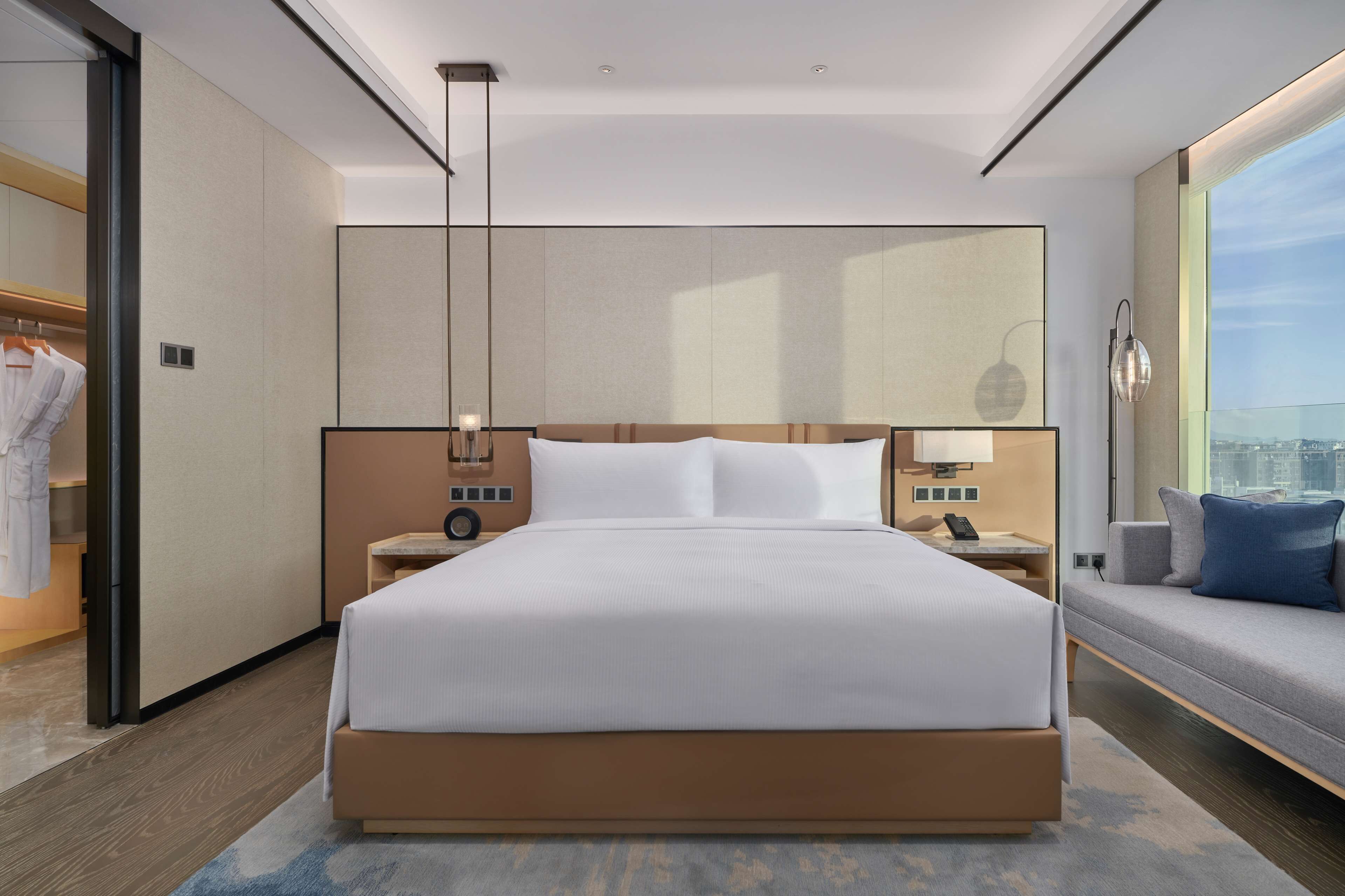 A modern hotel room with a large bed, bedside tables, a nightstand, a lamp, and a view of the city.