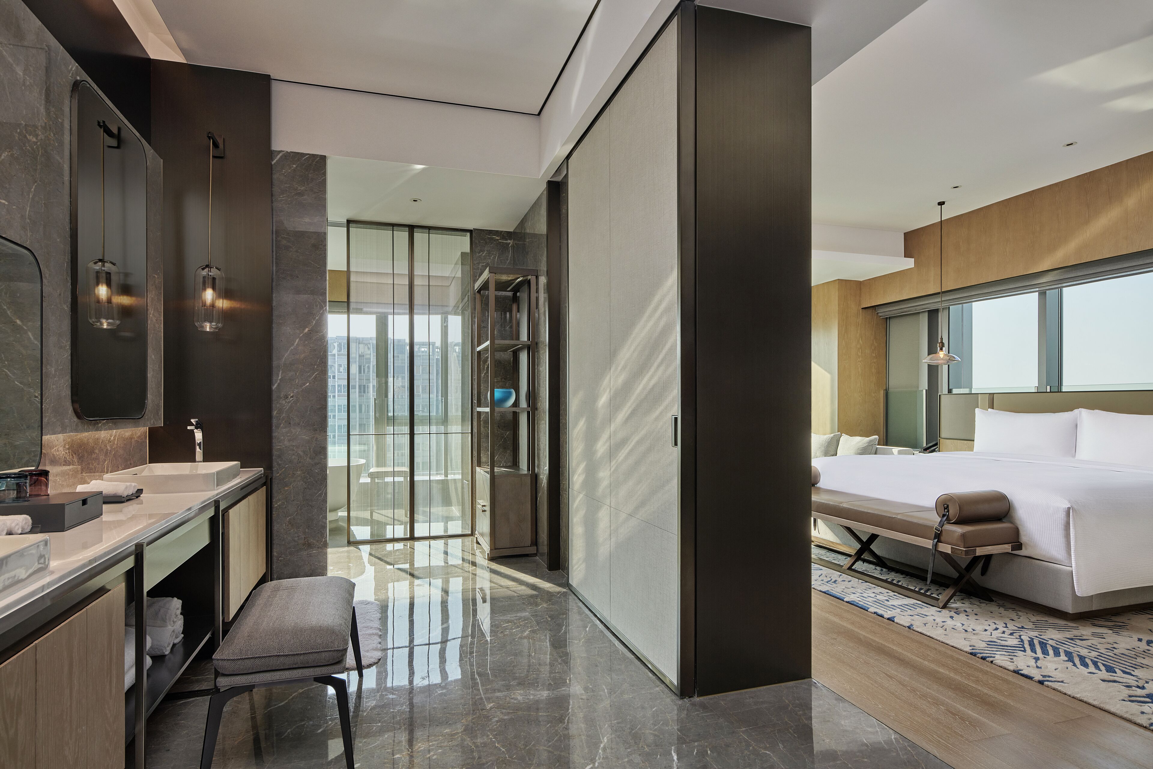 A modern hotel room with a large bed, a bathroom with a vanity and mirror, and a view of the city through a glass door.