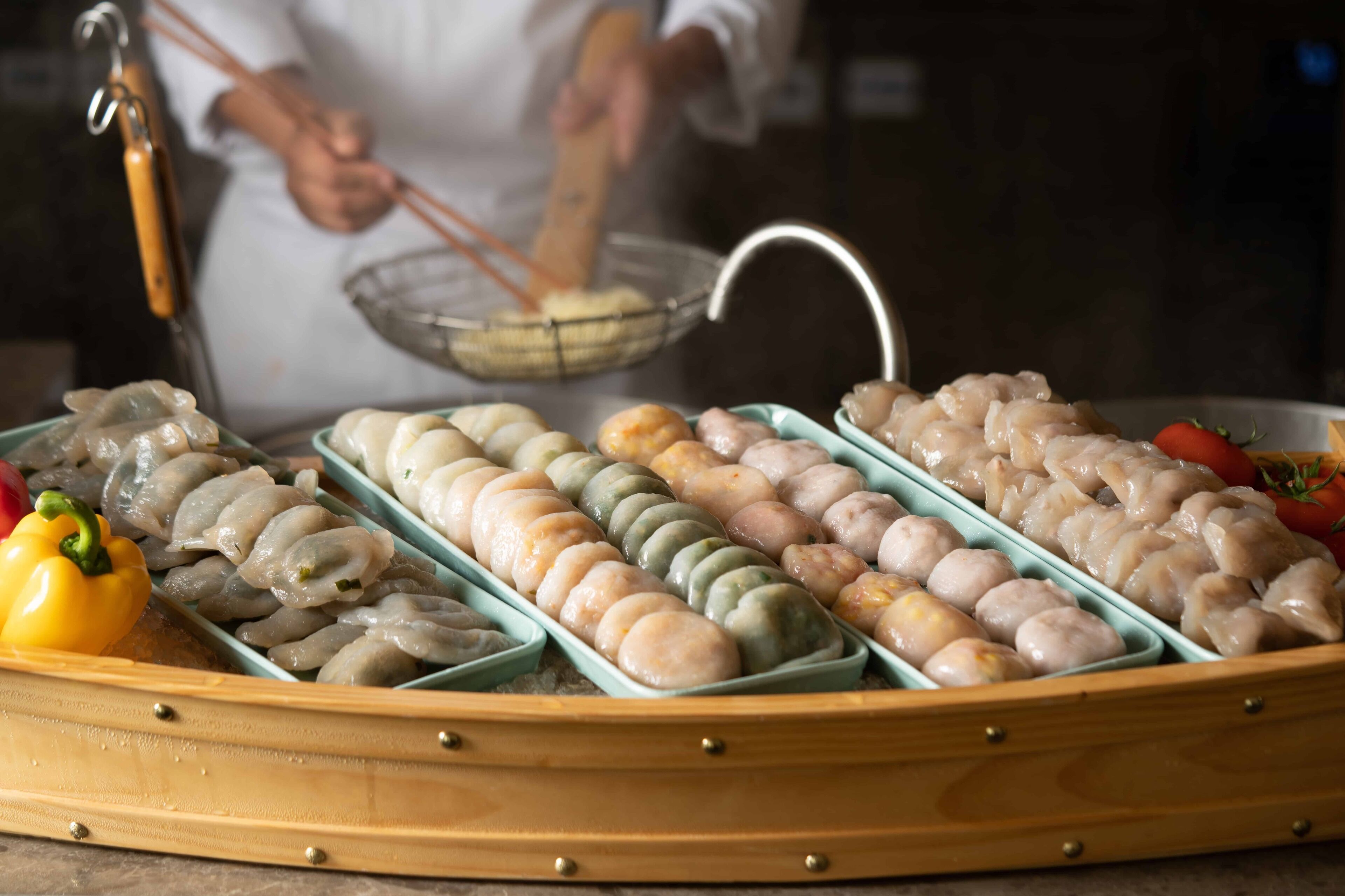 A variety of dumplings arranged in a wooden tray, with a chef in the background.