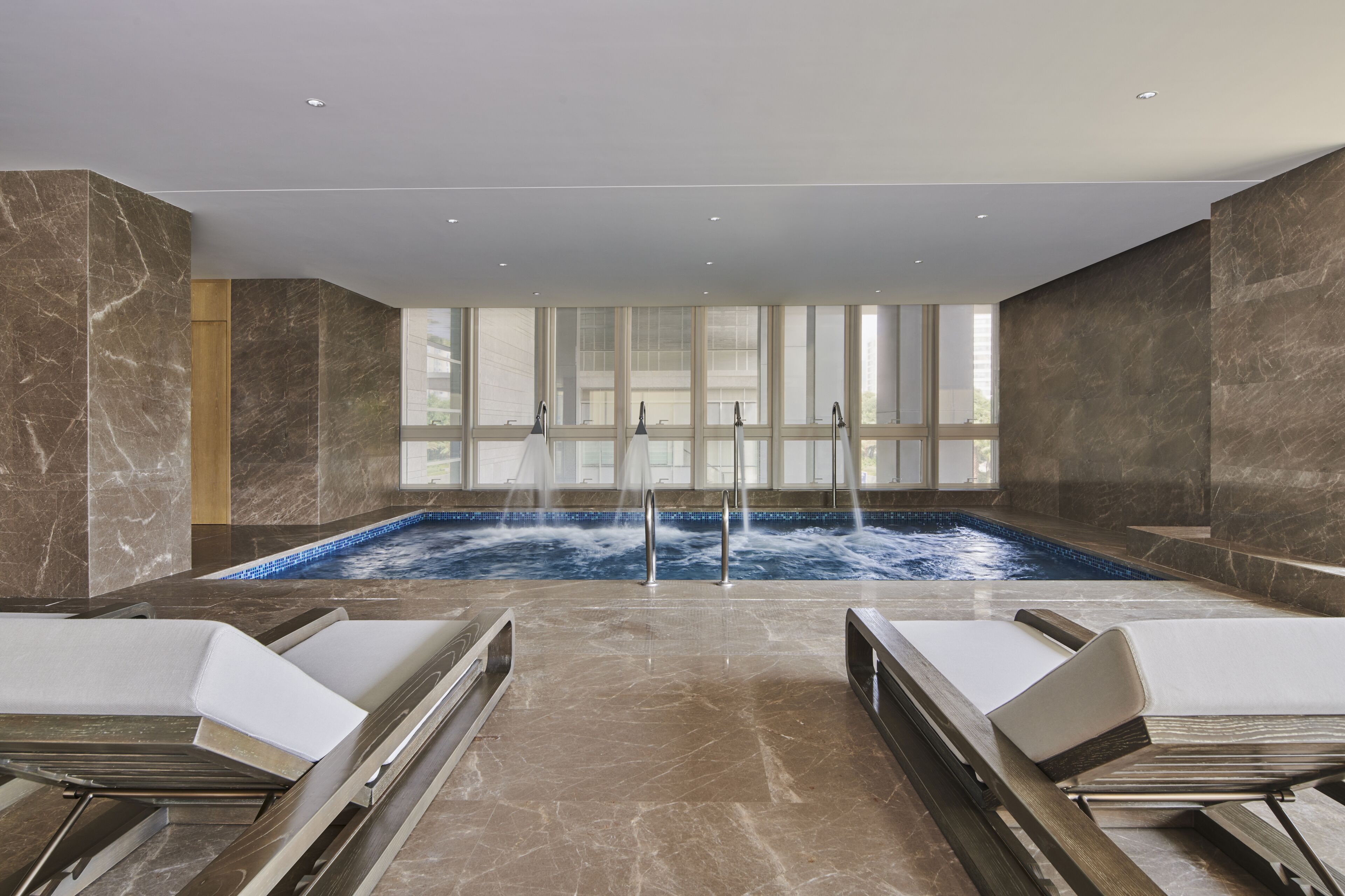 A spa area with a large indoor pool, two lounge chairs, and a view of the outside through large windows.