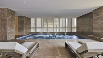 A spa area with a large indoor pool, two lounge chairs, and a view of the outside through large windows.