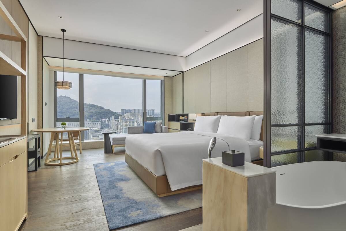 Ett modernt hotellrum med en stor säng, en sittgrupp, utsikt över staden och ett stort fönster.