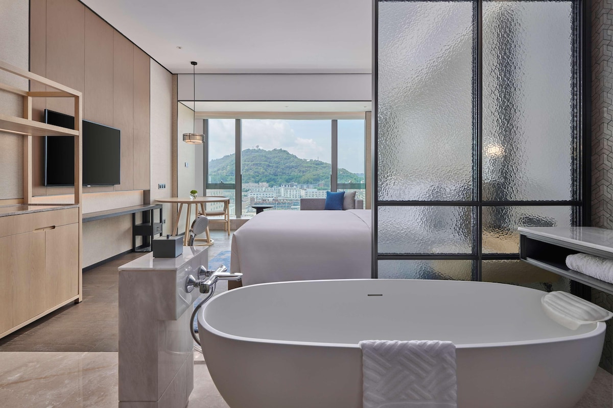 A modern hotel room with a large bathtub, a television, and a view of a cityscape through a large window.