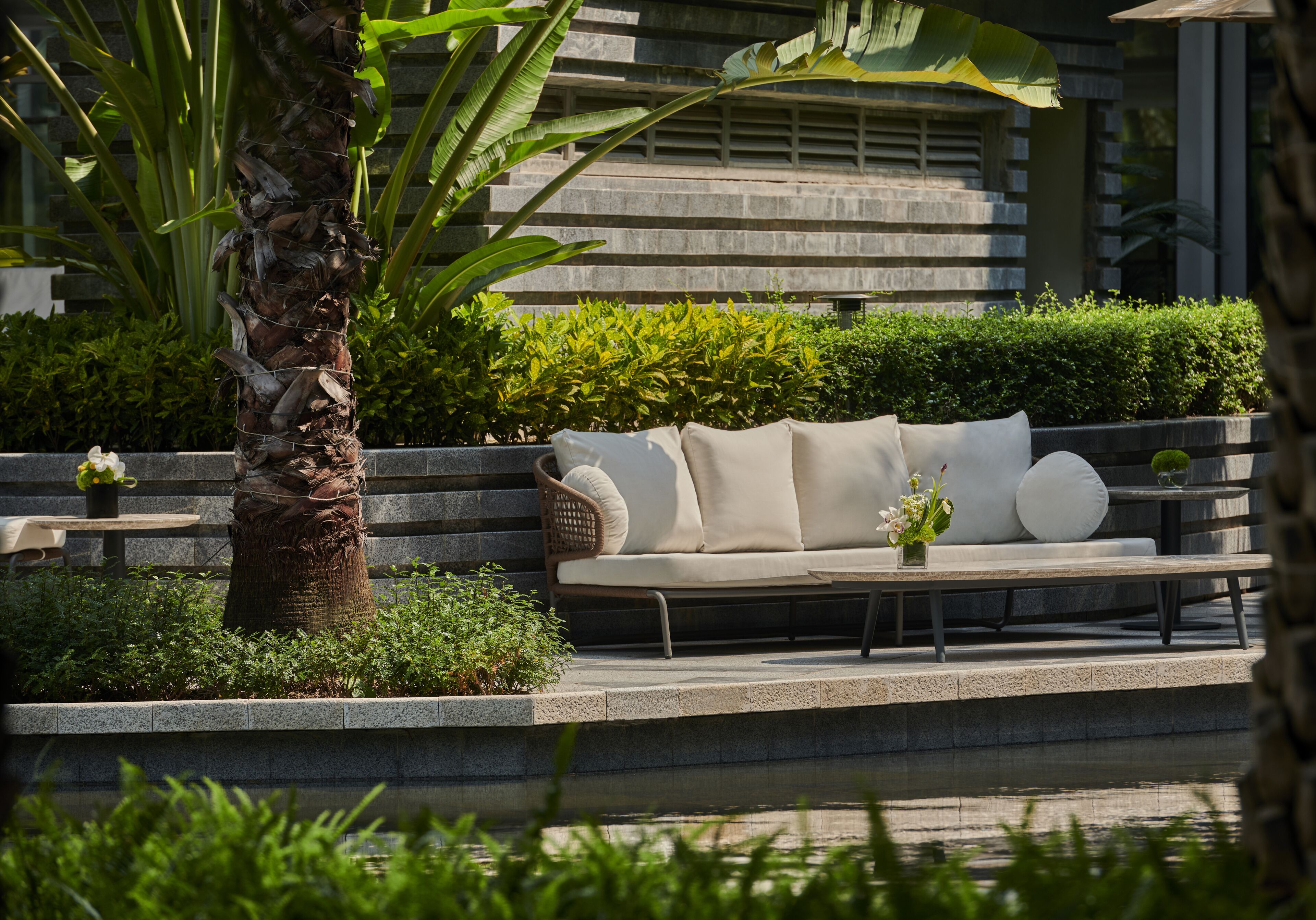 A modern outdoor seating area with a sofa and coffee table, surrounded by lush greenery and a water feature.