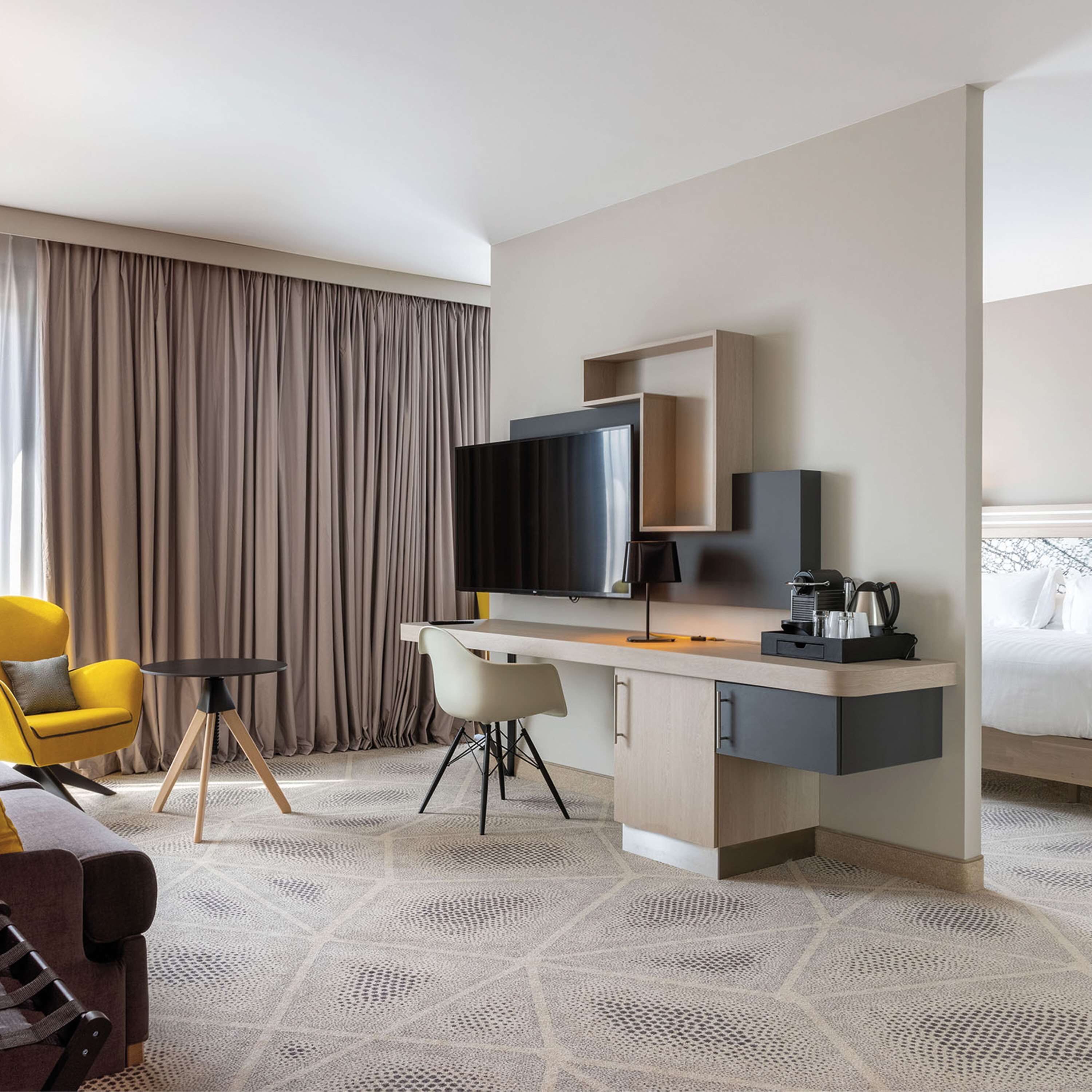 A modern hotel room with a flat-screen TV, a desk with a coffee maker, a yellow armchair, and a bed with white linens.