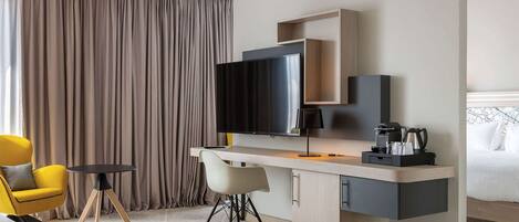 A modern hotel room with a flat-screen TV, a desk with a coffee maker, a yellow armchair, and a bed with white linens.